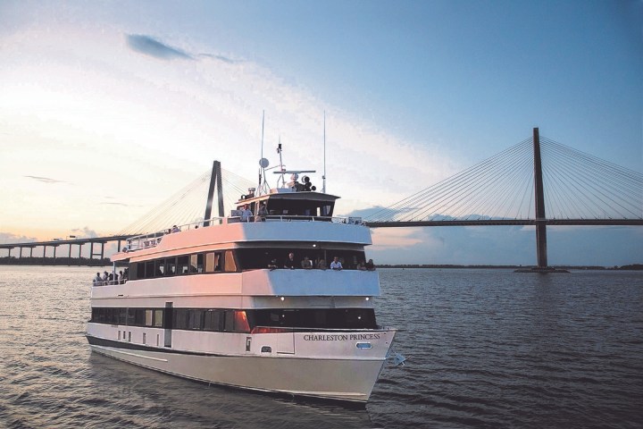 White cruise boat on river near a large suspension bridge at sunset.