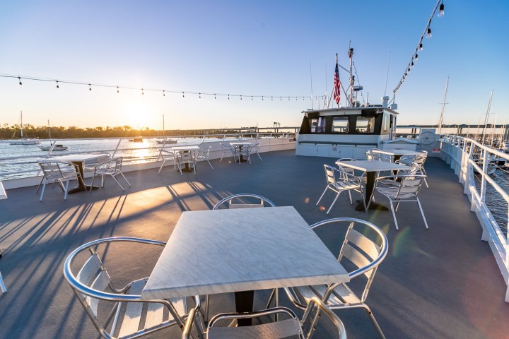 Sunlit boat deck with empty tables and string lights overlooking water.