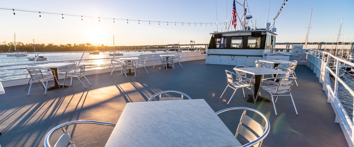 Sunlit boat deck with empty tables and string lights overlooking water.
