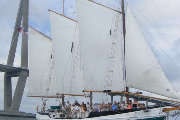 Sailing ship with white sails near a large suspension bridge.