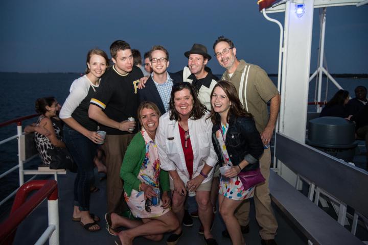 Group of eight adults smiling on a boat deck at dusk.