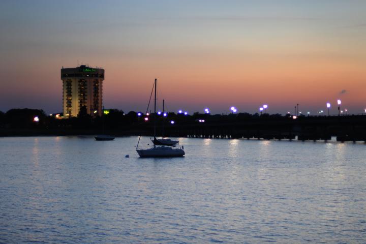 Sunset over water with a sailboat, distant building, and city lights.