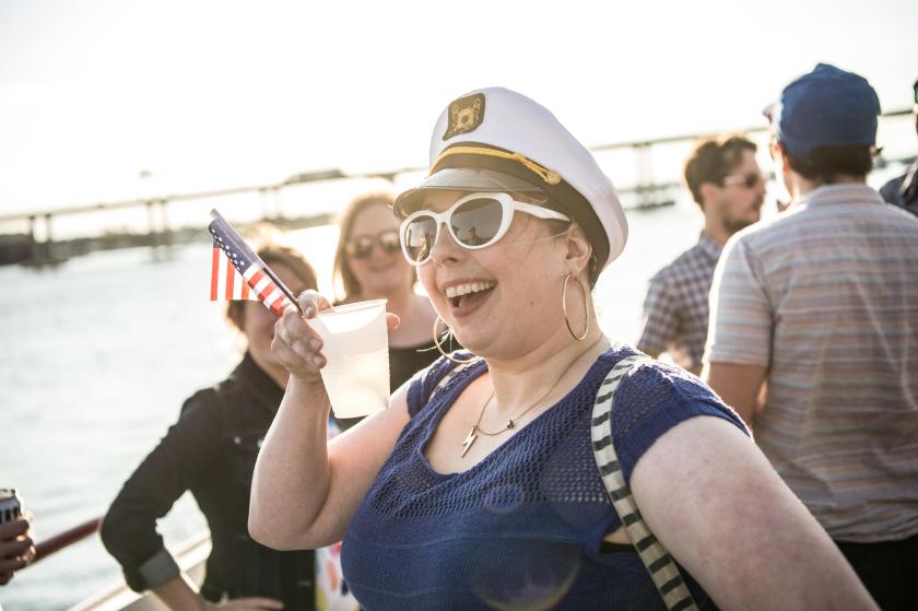 Person in captain's hat and sunglasses smiling, holding drink and small flag at outdoor gathering.