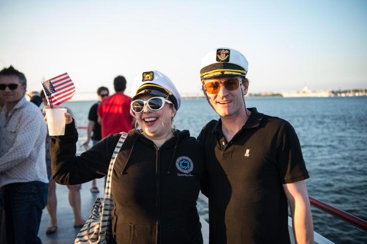 People in captain hats pose by water, one holding U.S. flag.