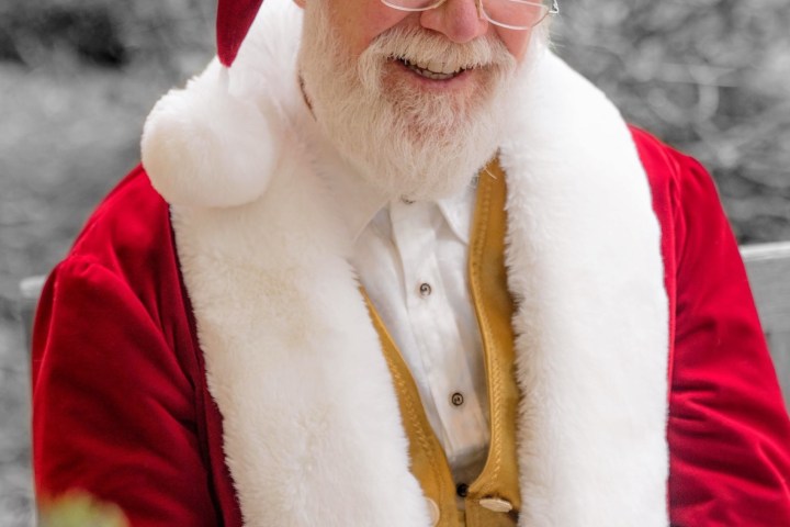Smiling man in Santa costume with white beard and glasses sitting outdoors.
