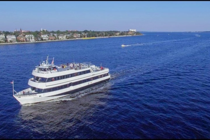 Large white yacht sailing on a wide river with a shoreline in the background.