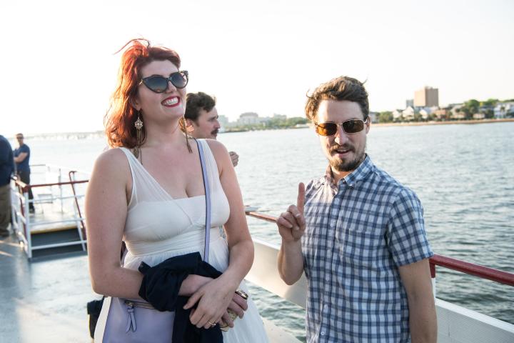 Two people smiling on a boat with water and cityscape in the background.