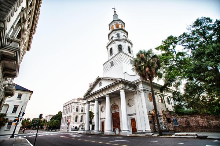 White church with a tall steeple on a city street lined with trees.