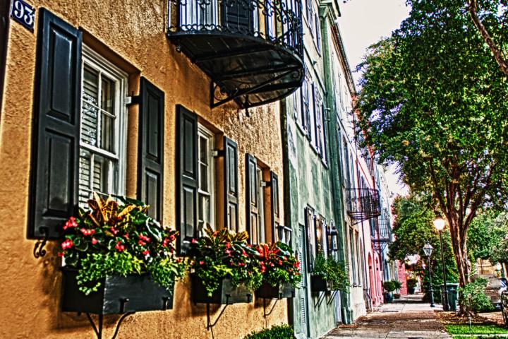 Colorful row of historic houses with balconies and flower boxes on a tree-lined street.