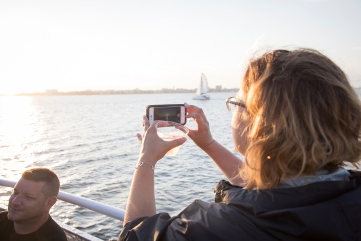 Person on a boat photographing a sailboat with their phone at sunset.