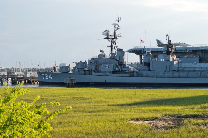 Grey naval ship docked near lush grassy shore.