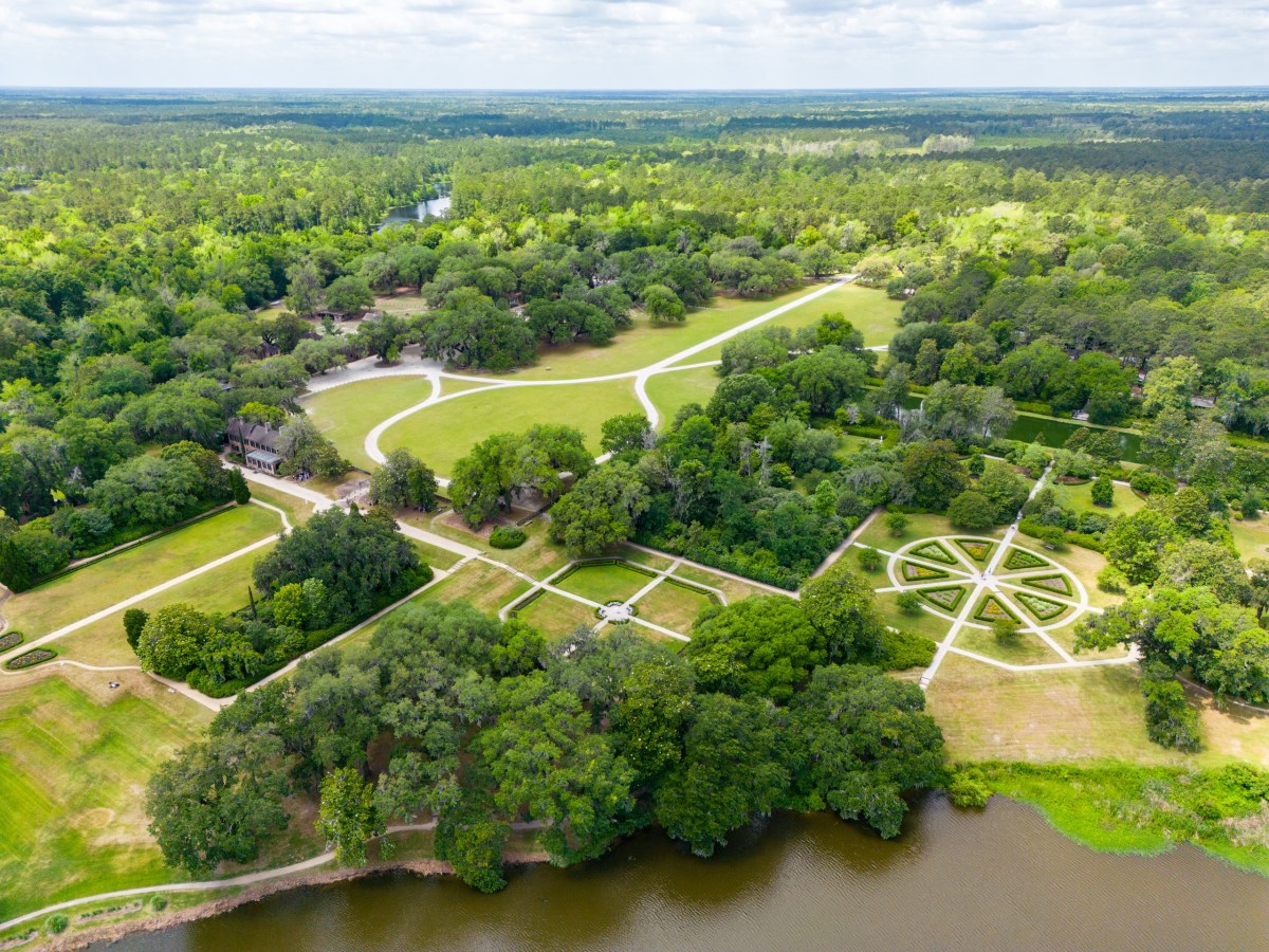 Aerial view of large estate with gardens, lawns, and pathways surrounded by trees near a river.