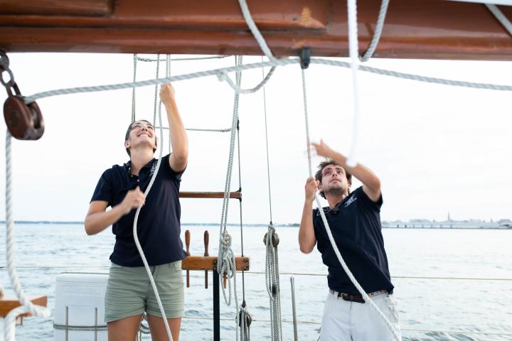 Two people adjusting ropes on a sailboat, with a sea backdrop.