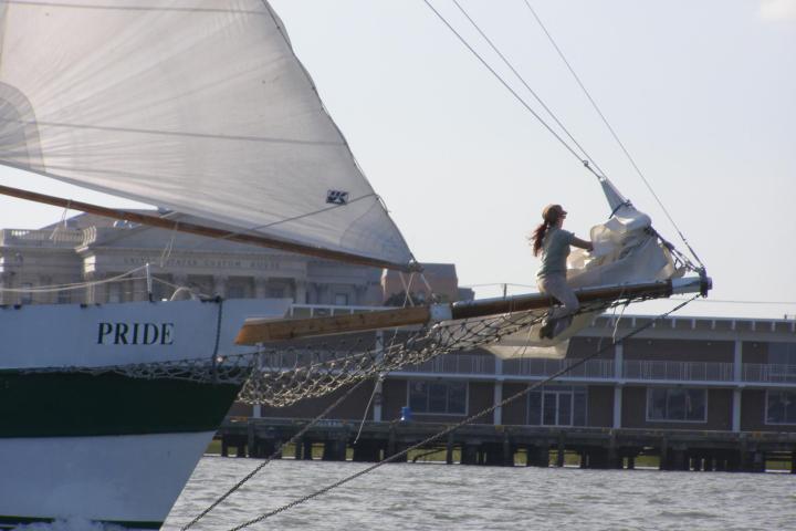 Person on bow of sailboat named Pride in water, building in background.