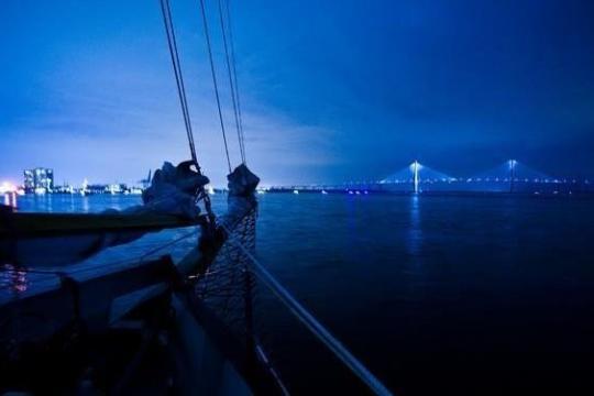 Sailboat at night, city lights and bridge in the distance.