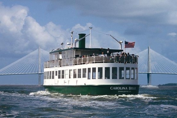 Ferry boat 'Carolina Belle' on water, with U.S. flag, bridge, and cloudy sky.