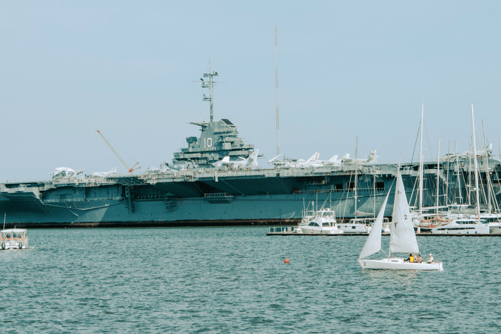 Large aircraft carrier docked near sailboats on a calm sea.