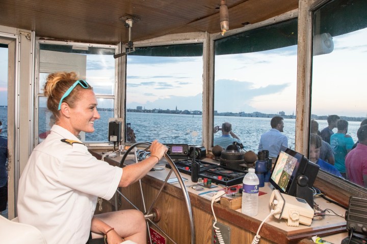 Smiling ferry captain steering with cityscape in view.