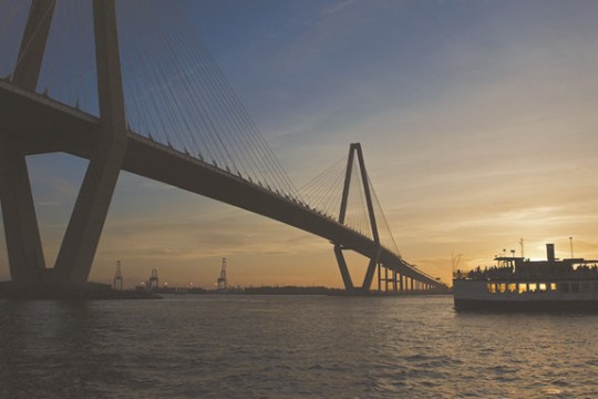 Sunset view of a bridge over water with a ferry sailing underneath.