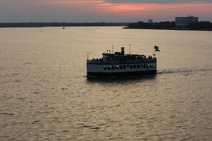 Ferry on a calm river at sunset with buildings in the distance.