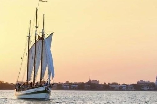 Sailboat on water with city skyline and sunset in background.