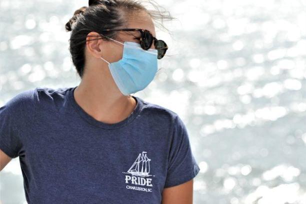 Person with sunglasses and mask gazes sideways, wearing a Pride shirt near shimmering water.