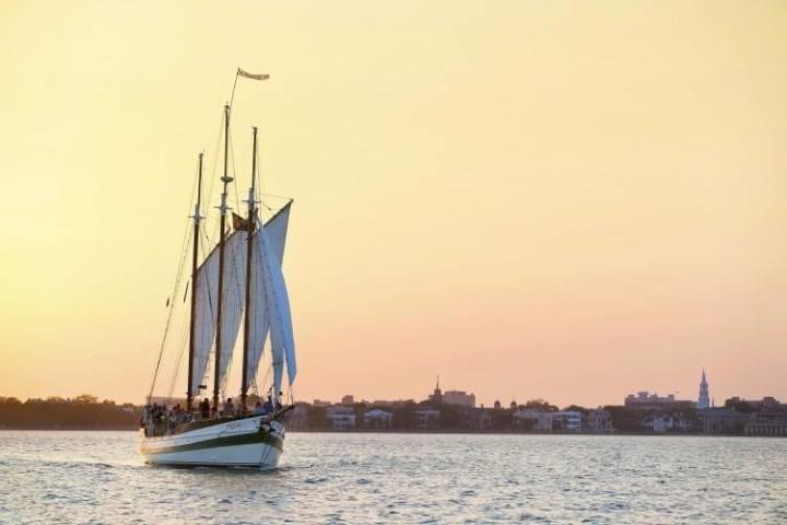 Sailing ship on calm water at sunset with city skyline in background.
