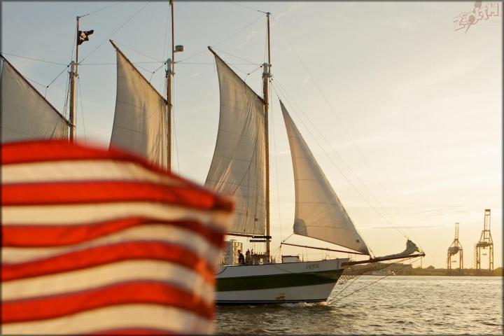Sailing ship with three masts near a waving American flag at sunset.