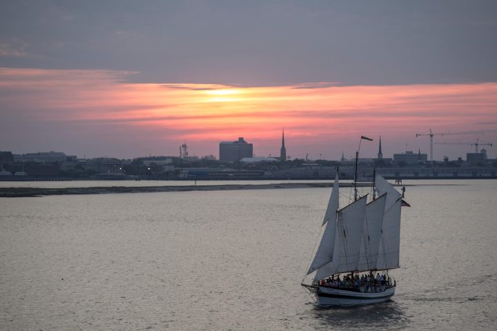 Sailboat on calm water at sunset with city skyline in background.