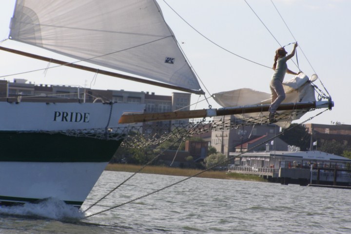 Person standing on a ship's bowsprit over water with buildings in the background.