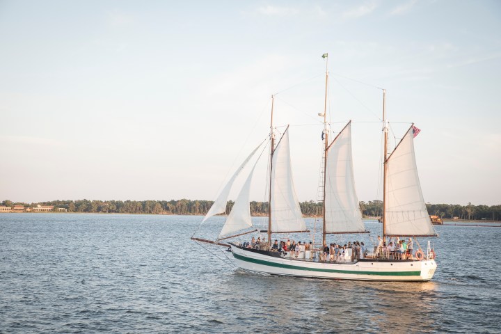 Sailboat with three sails on a calm lake, people on deck, forest in background.