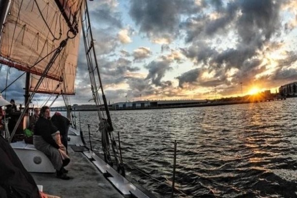 People on a sailboat at sunset, with clouds and water reflecting light.