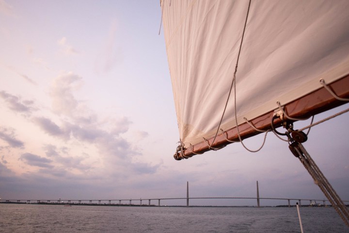 Sailboat on water with a bridge and cloudy sky at sunset.