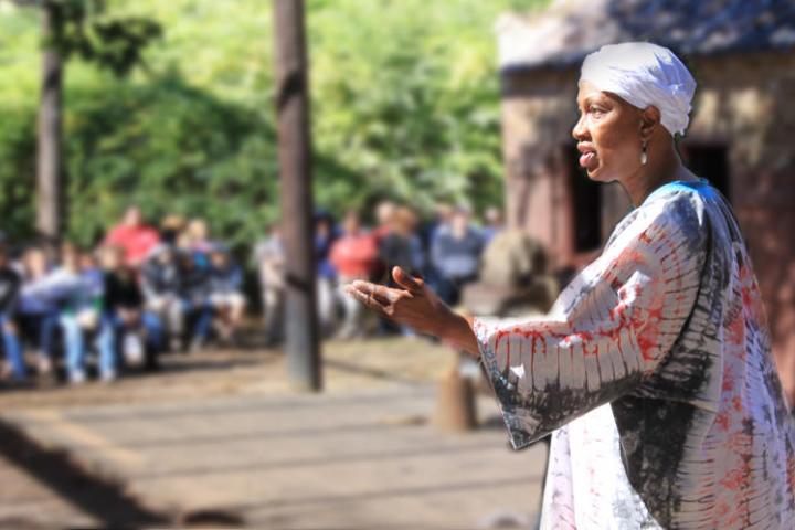 Woman in a white headscarf speaks outdoors to a seated audience.