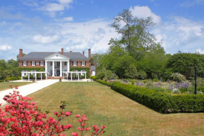 Large brick house with columns, garden, and driveway under a blue sky.