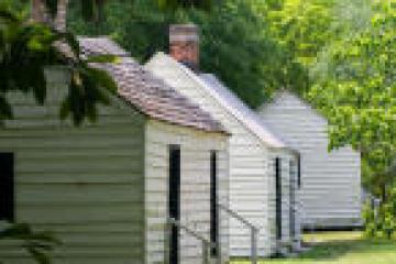 Row of small, white cabins with green shutters surrounded by trees on a sunny day.